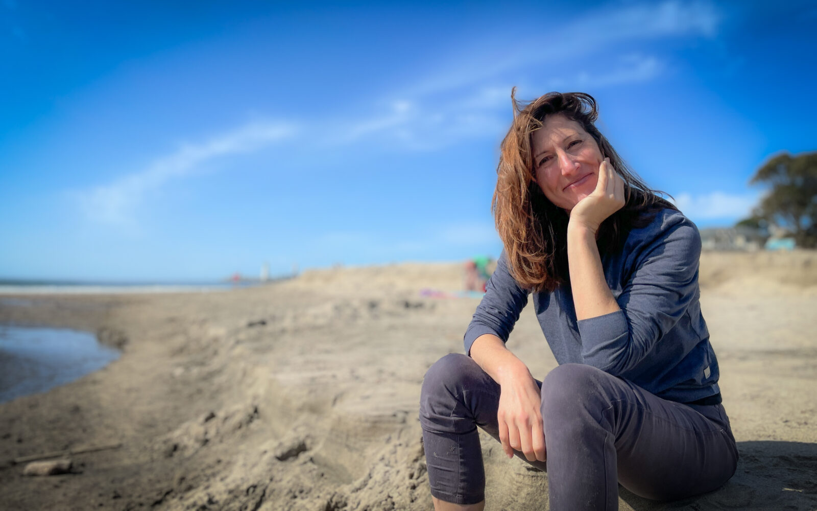 Lindsey Dillon sits on a sandy beach, chin resting in her hand, smiling warmly at the camera. Her auburn hair is windswept across her face. The light is golden and coastal, late afternoon, the kind of California light that makes everything look like it was always supposed to be exactly this way.