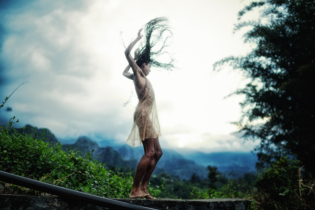 Adji Cissoko of Alonzo King LINES Ballet in a light-colored dress stands on a hillside path with long braided hair whipping dramatically through the air, silhouetted against a misty mountain landscape with dramatic cloudy skies.