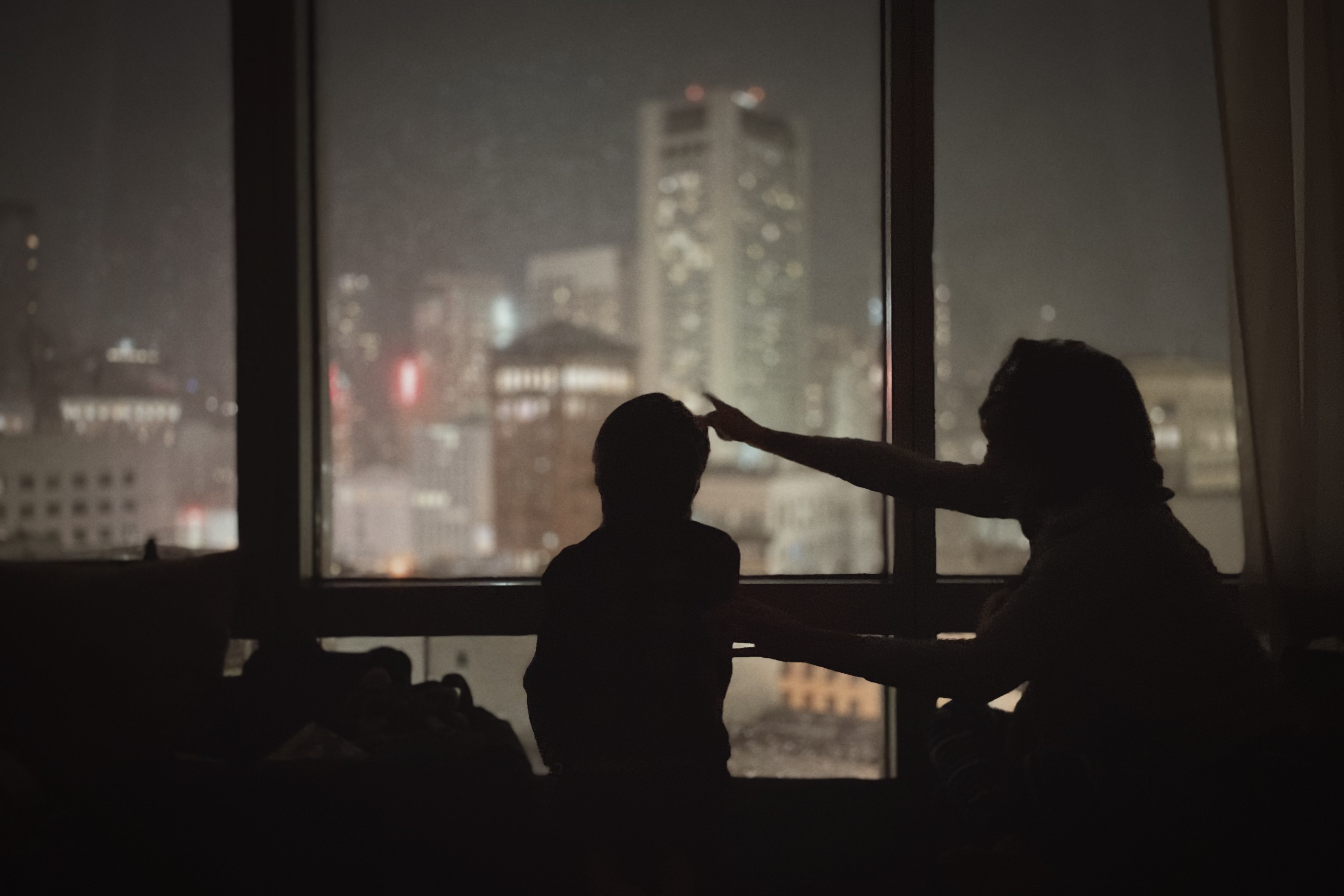 Silhouettes of mother and child, Lindsey Dillon and Charlie Lyons,in a darkened room, one appearing to touch or adjust the other's head, framed against a large window overlooking a misty cityscape of illuminated buildings at night at the Four Seasons Hotel, San Francisco.